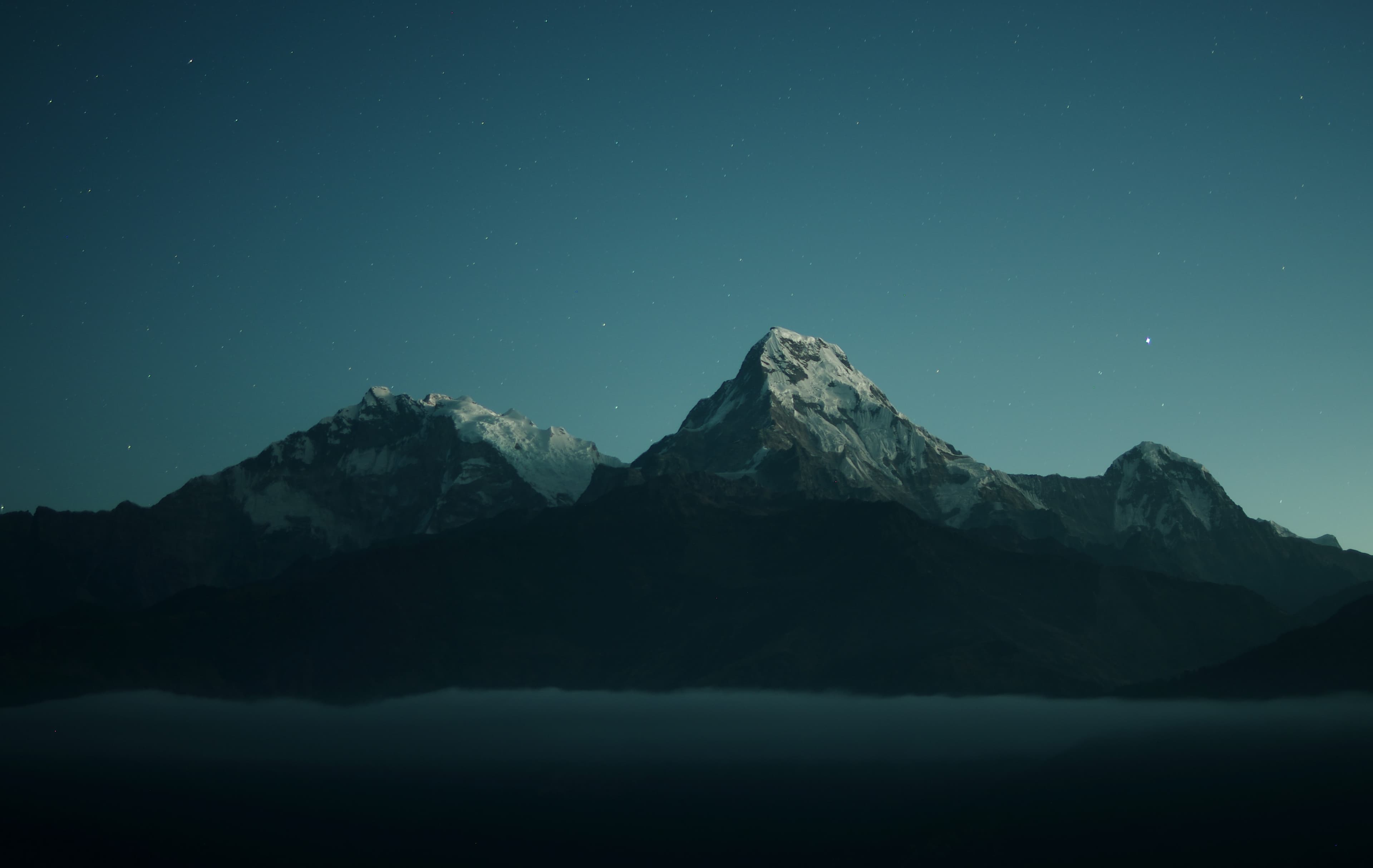 Photograph of snowy large mountains in the distance, clear starry blue night sky behind them